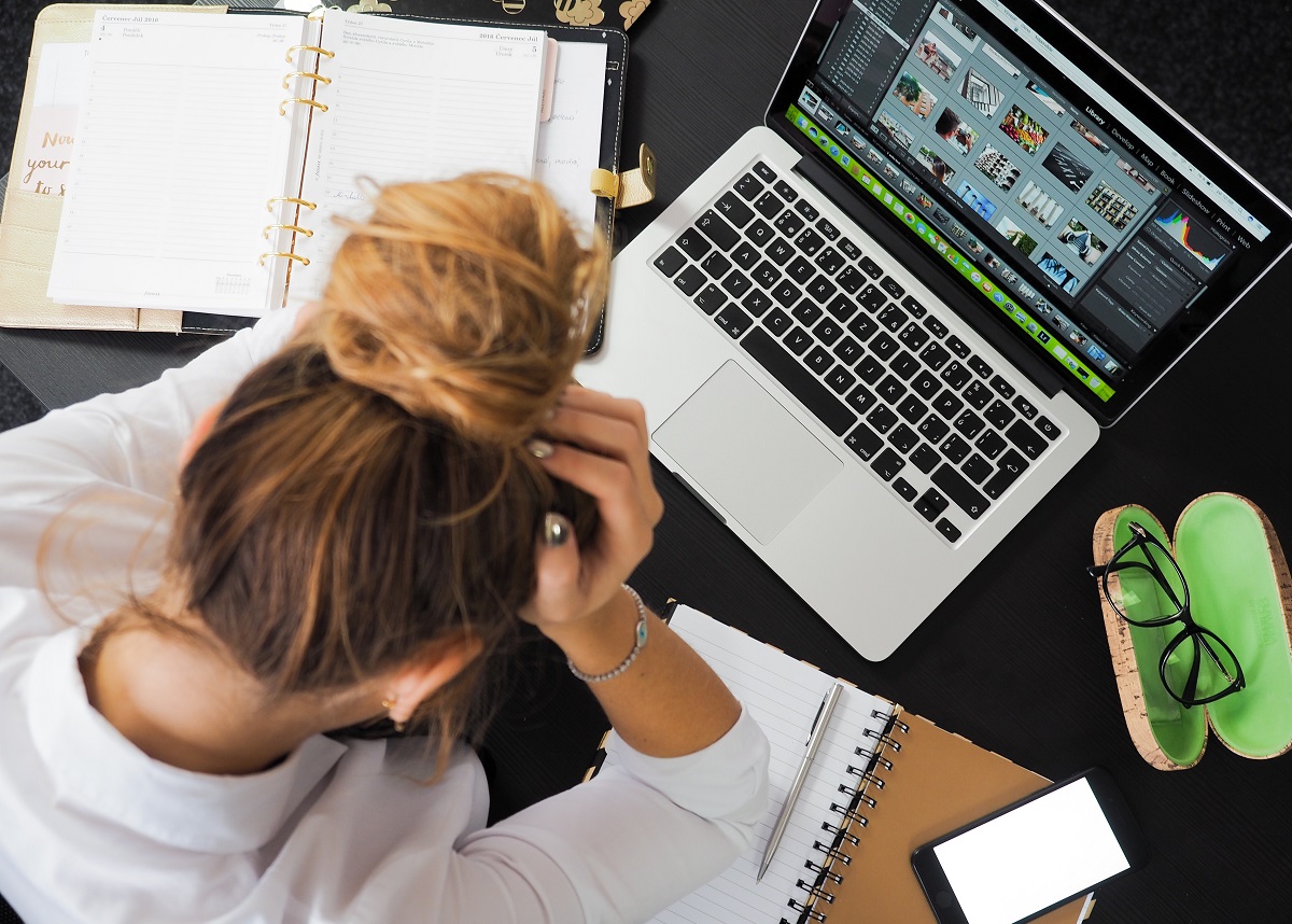 Mulher com a mão na cabeça e notebook, caderno, óculos, celular espalhados ao redor