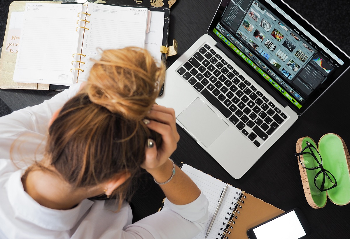 Mulher com as mãos na testa e em frente ao notebook e cadernos espalhados