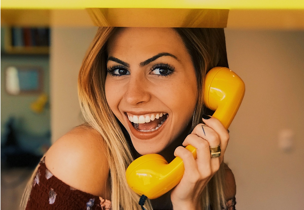 Mulher loira sorrindo e segurando um telefone amarelo na mão