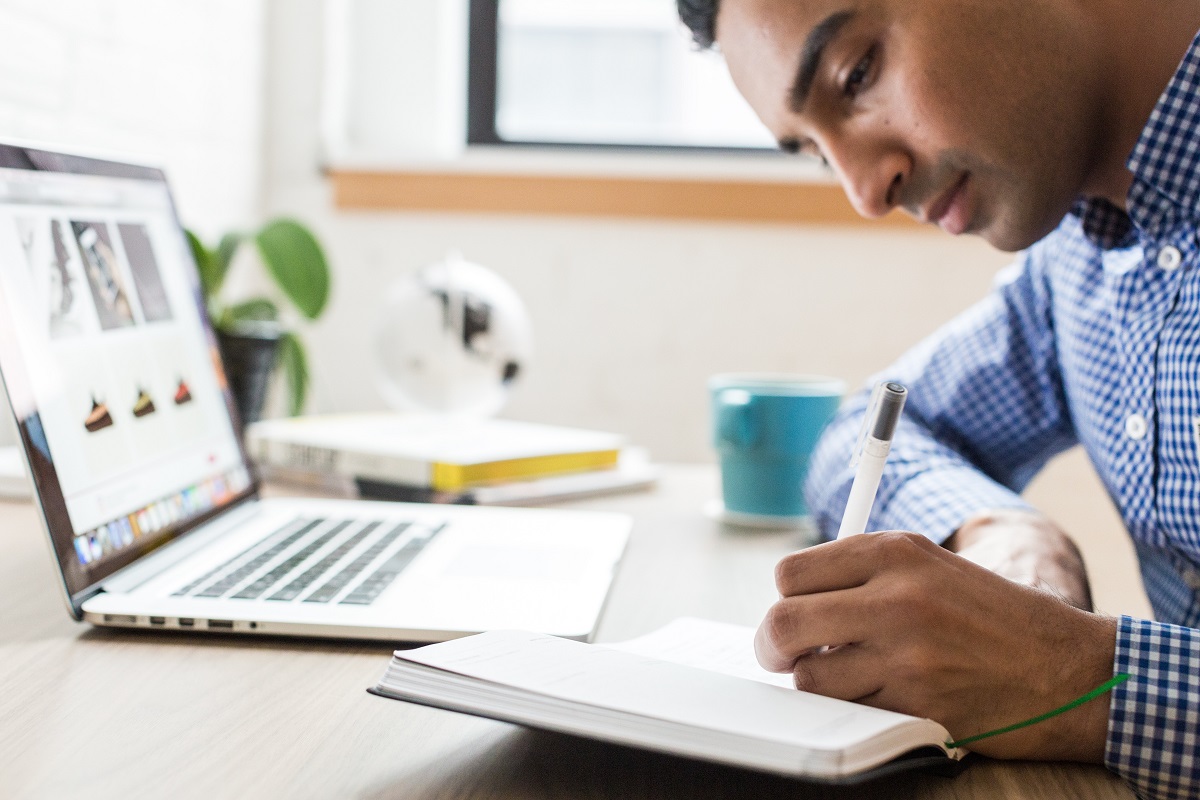 Homem sentado em uma cadeira em frente ao notebook, escrevendo em um caderno