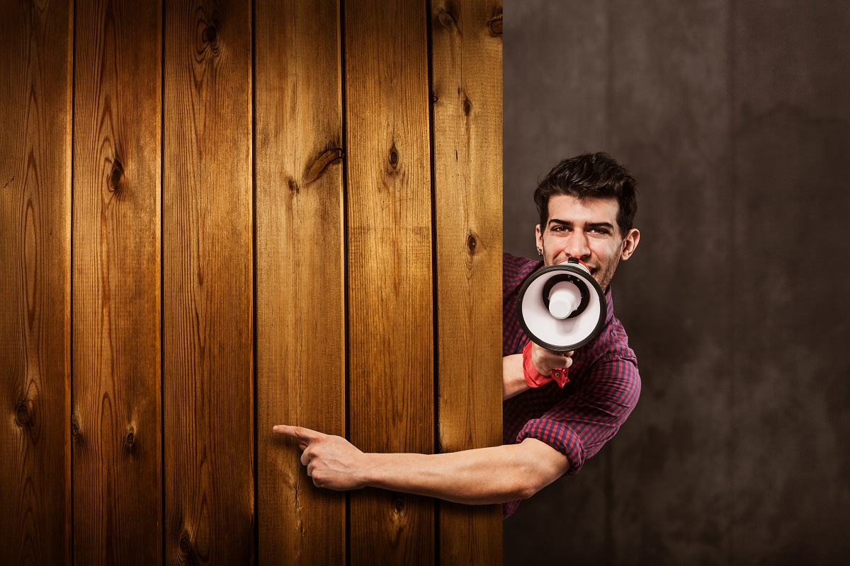 Homem com um megafone na mão e apontando para um muro de madeira com a outra mão