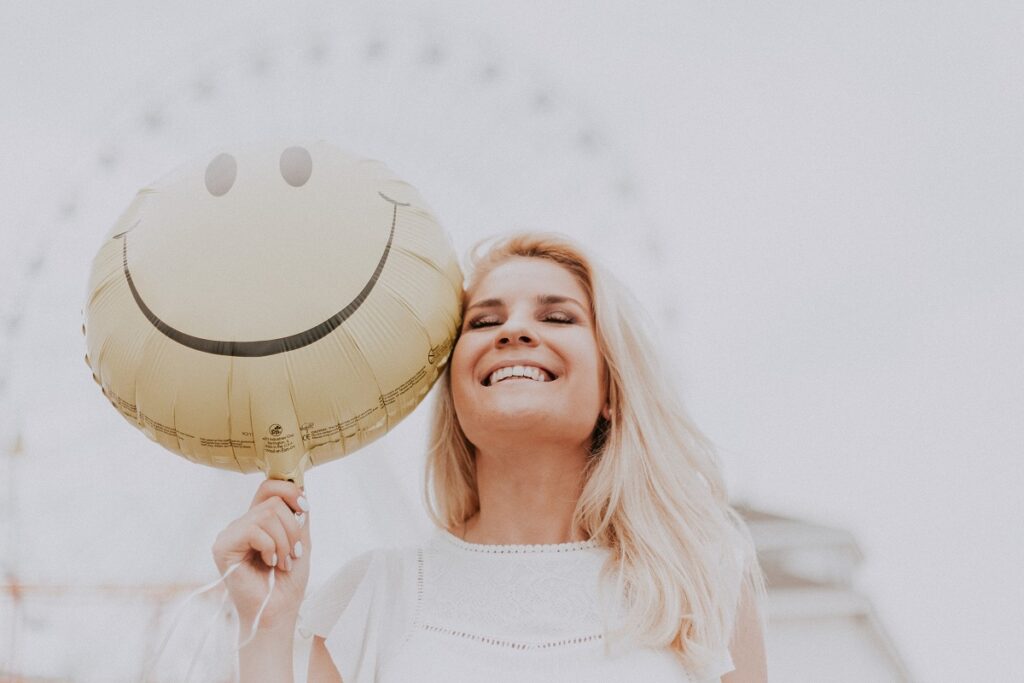 Pessoa sorrindo segurando um balão com carinha sorrindo ao lado do rosto