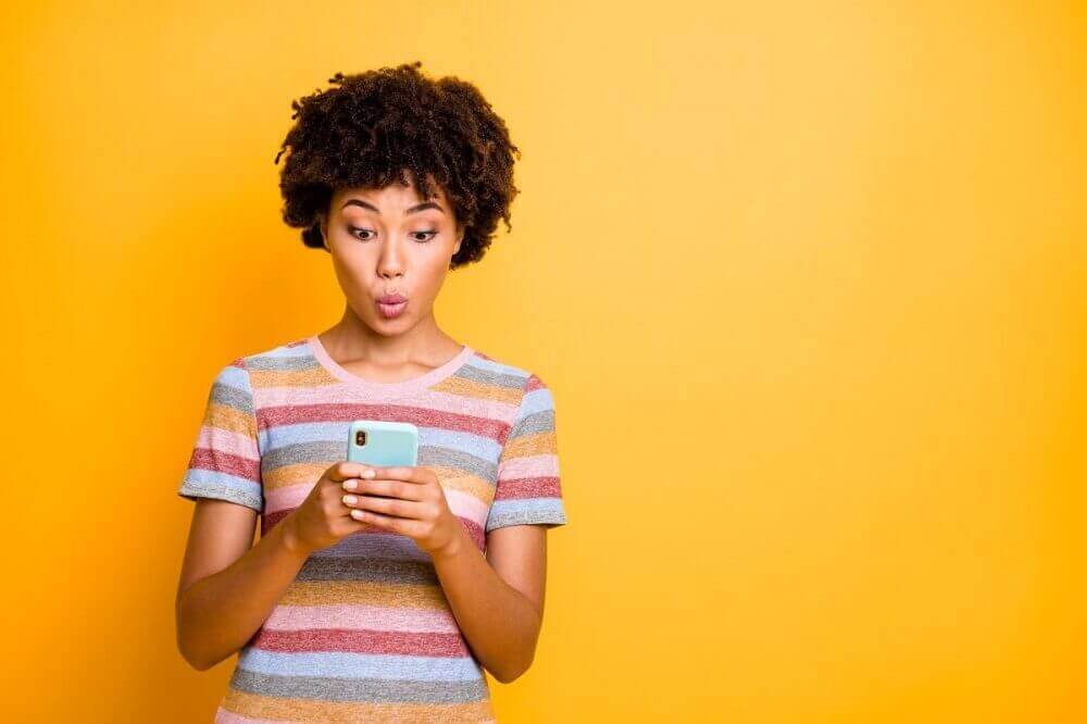 Mulher negra de cabelos crespos e curtos, camiseta colorida, segurando um celular azul em suas mãos. O fundo da foto é laranja.