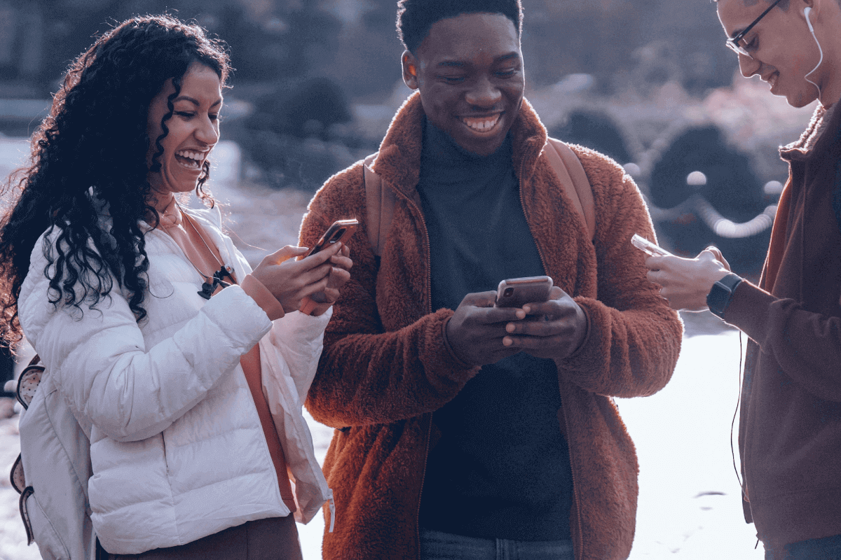 Mulher à esquerda, homem ao centro e à direita, todos eles segurando um celular em suas mãos, olhando para a tela e sorrindo