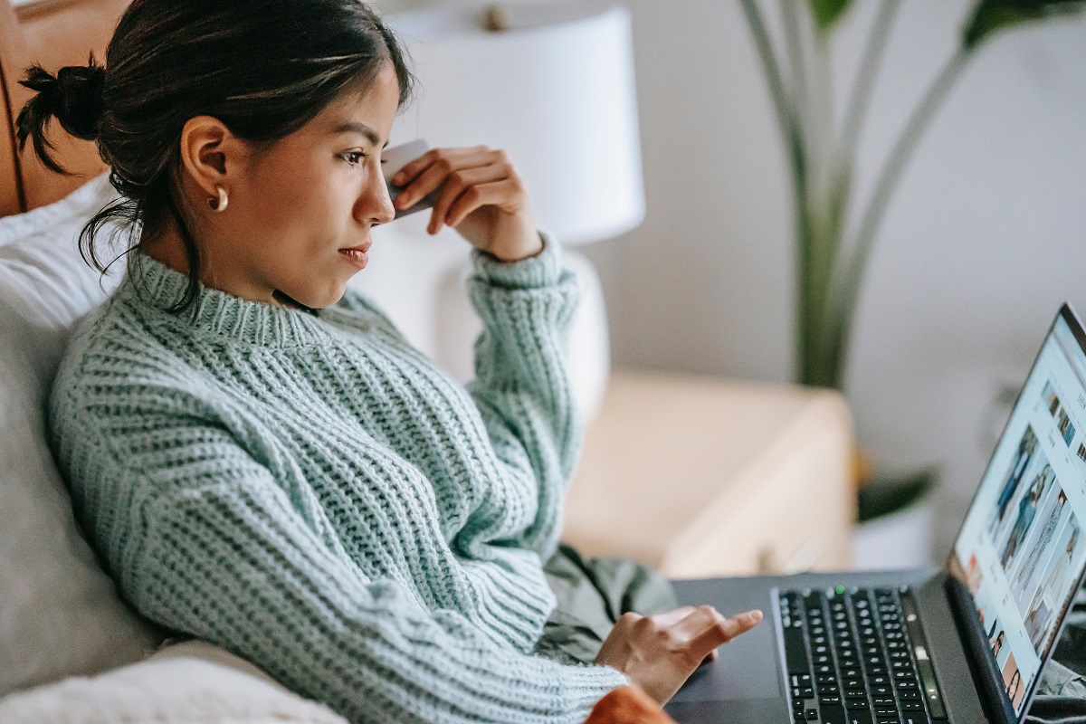 Mulher com blusa de lã em frente a um notebook olhando para uma página de compra e com um cartão de crédito na mão esquerda