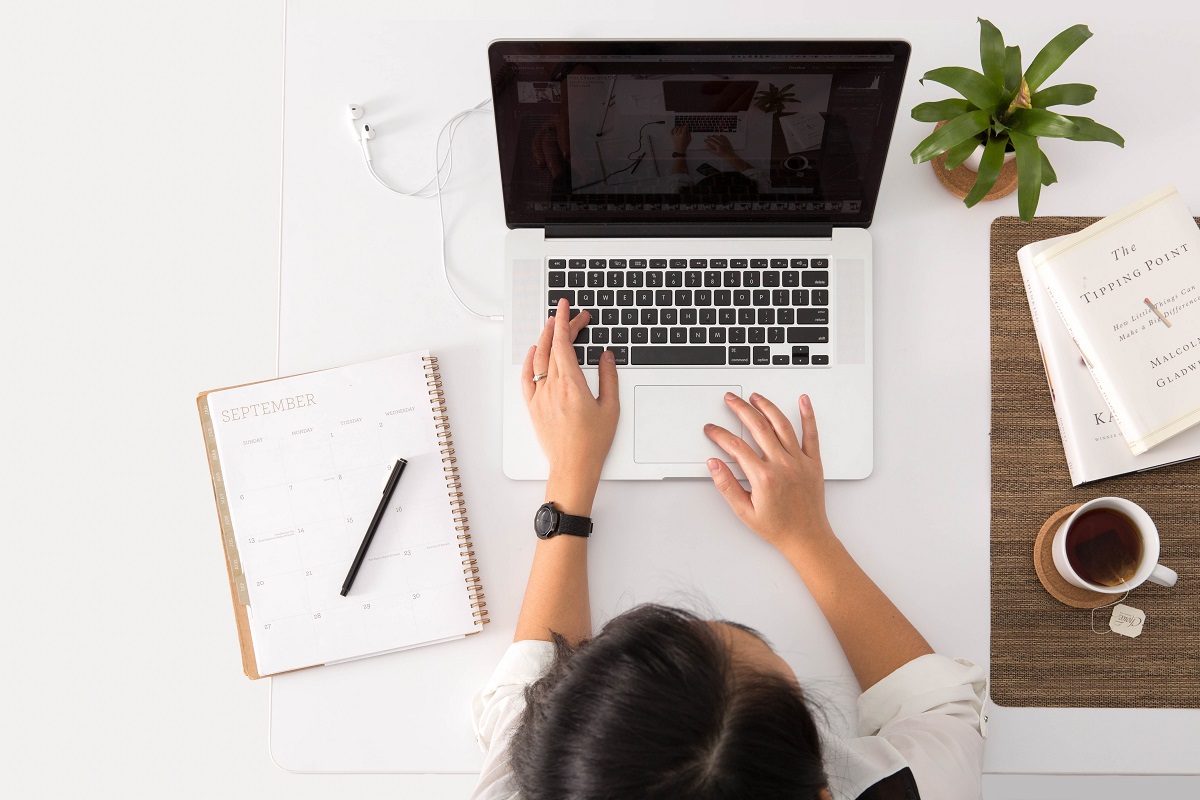 pessoa digitando algo em seu notebook; em cima da mesa existe uma agenda, livro, xícara com café e uma pequena planta