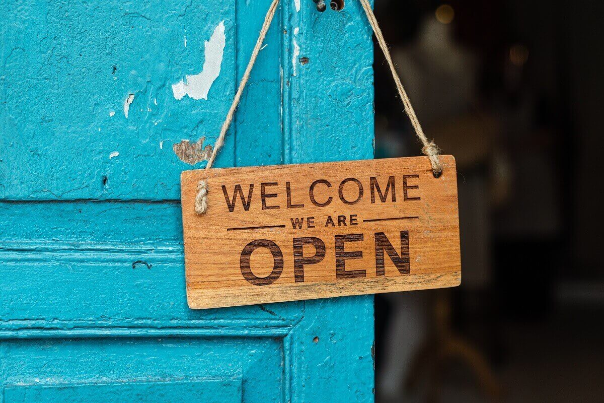 Porta azul com uma placa em madeira escrito "Welcome we are open", que em português é "Boas-vindas, estamos abertos"